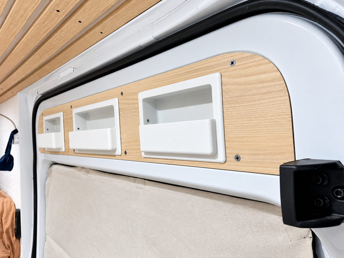 Close-up of a Mercedes Sprinter van interior showing light wood paneling with three recessed white storage compartments, a white door frame, and part of a beige cushion or seat below.