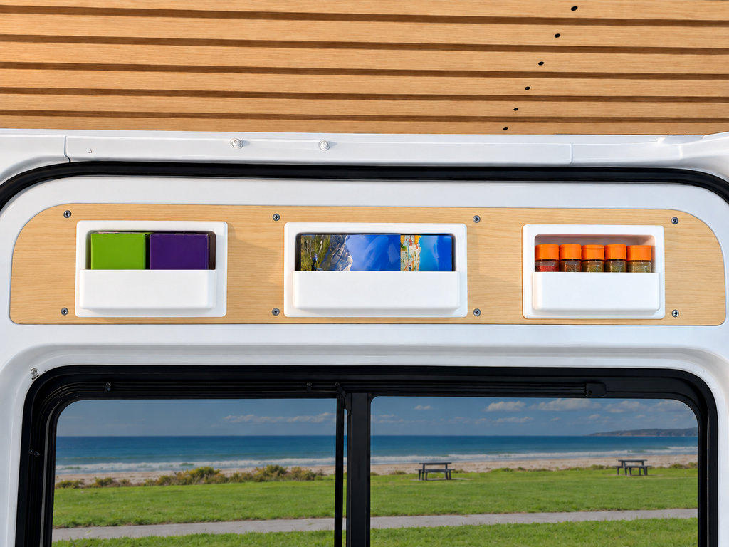 Close-up of a Sprinter Camper interior window trim with three small rectangular storage boxes from Barn House Studio, above a large window showing an outdoor beach and grassy landscape.