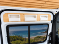 A close-up of a white Mercedes Sprinter camper van interior showing three recessed white storage compartments, from Barn House Studio, mounted on a light wood panel above a window with a coastal ocean view reflected inside.