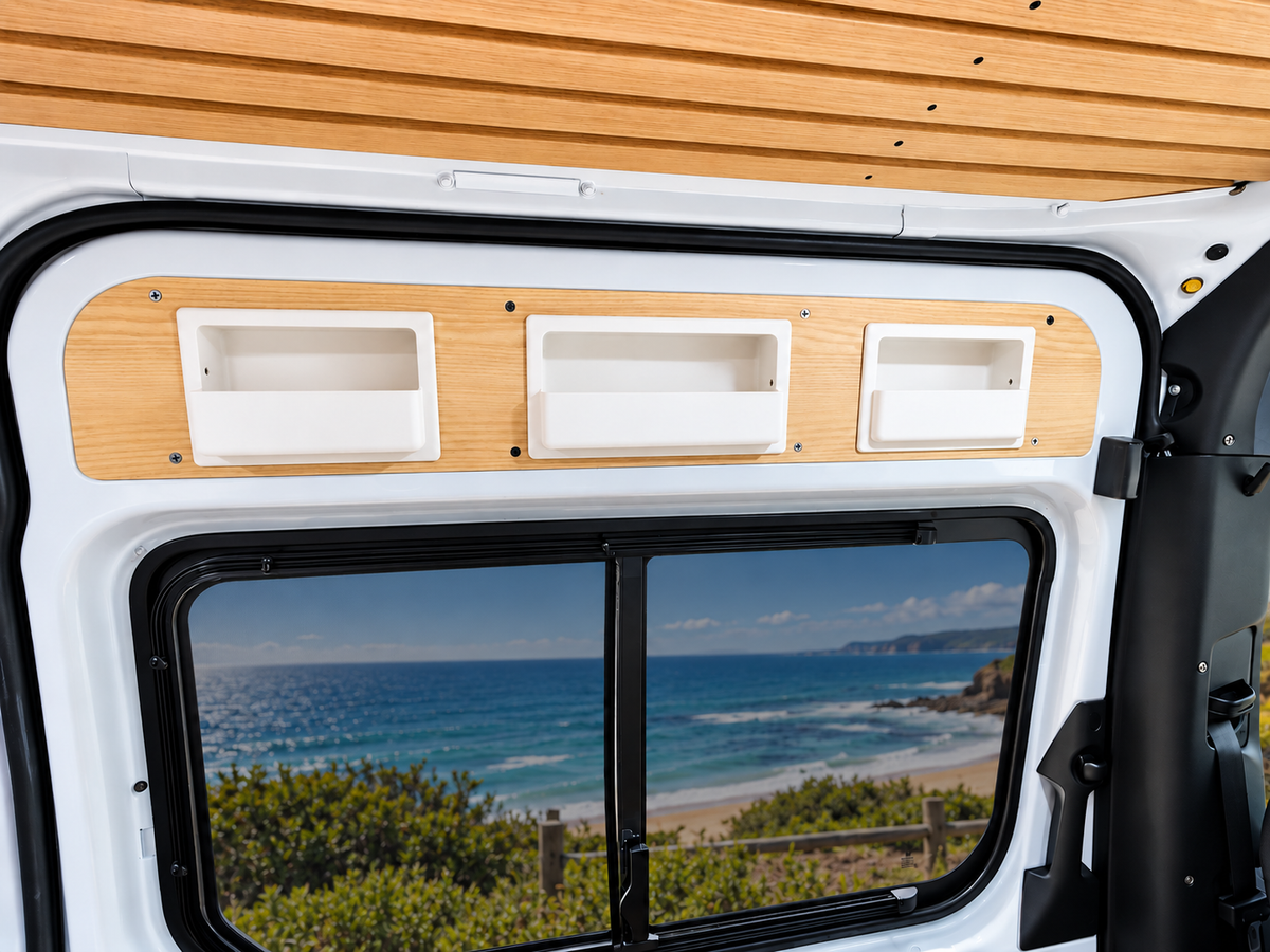 A close-up of a white Mercedes Sprinter camper van interior showing three recessed white storage compartments, from Barn House Studio, mounted on a light wood panel above a window with a coastal ocean view reflected inside.
