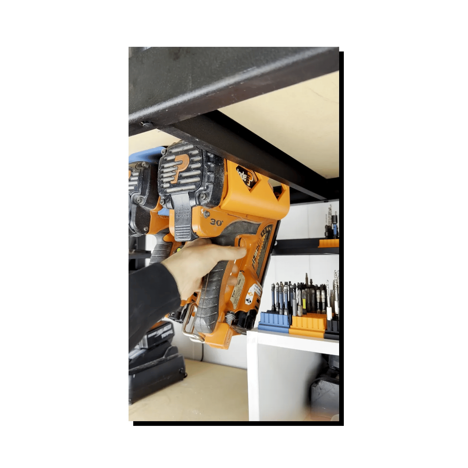 Hand holding a nail gun in a large tool holster in a workshop setting with tools and equipment.